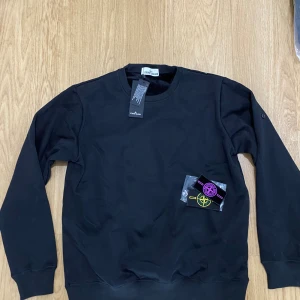 Svart sweatshirt från Stone Island - Svart sweatshirt från Stone Island med klassisk rund hals och ribbade muddar. Medföljer två utbytbara Stone Island-patchar i lila och gul. Tröjan är tillverkad i mjuk bomull och har långärmad passform, perfekt för en clean och stilren look.