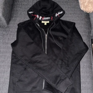 Svart hoodie från Burberry med dragkedja - Svart hoodie från Burberry med klassiskt rutigt foder i huvan och broderad logga på bröstet. Tröjan har hel dragkedja, snörning i huvan och kängurufickor. Perfekt för dig som gillar stilren design med en touch av lyx.