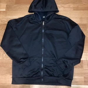 Svart hoodie med dragkedja - Svart hoodie med hel dragkedja framtill och huva. Tröjan har två fickor på magen och ribbade muddar vid ärmslut och nederkant. Materialet känns mjukt och är troligtvis polyester eller bomull. Perfekt för chill dagar eller när du vill ha något enkelt och bekvämt.