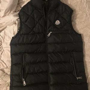 Svart dunväst från Moncler - Svart dunväst från Moncler med quiltad design och hög krage. Västen har dragkedja framtill, två sidofickor med dragkedja och en bröstficka med dragkedja. Moncler-logga på bröstet och diskret Moncler-tryck vid ärmöppningarna. Perfekt för lager-på-lager.