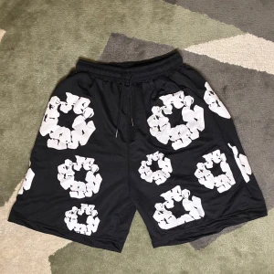 Svarta shorts från Denim Tears - Svarta shorts från Denim Tears med stora vita blommönster tryckta över hela plagget. Modellen har elastisk midja med snörning för skön passform och är tillverkad i mjukt material som passar perfekt för chill dagar. Snygg och unik design som sticker ut.