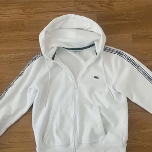 Vit hoodie från Lacoste med dragkedja - Vit hoodie från Lacoste i klassisk passform, med huva och snörning. Dragkedja framtill och två fickor. Svarta Lacoste-loggor längs ärmarna. Mjuk insida och ribbade muddar. Perfekt för chill eller streetwear.
