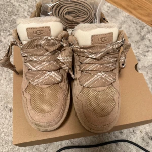 Beige sneakers från UGG med fårfoder - Snygga beige sneakers från UGG med rutiga snören och mjukt vitt fårfoder på insidan. Ovandelen är i mocka och textil, med detaljer i vitt och grov sula. Perfekta för höst och vinter när du vill ha både stil och värme.