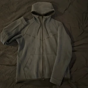 Mörkgrå hoodiejacka från Nike - Säljer en mörkgrå hoodiejacka från Nike med dragkedja hela vägen, huva med snörning och två stora fickor framtill. Jackan har en sportig look och är tillverkad i mjukt bomullsmaterial, perfekt för chill eller träning. Diskret Nike-logga på bröstet.