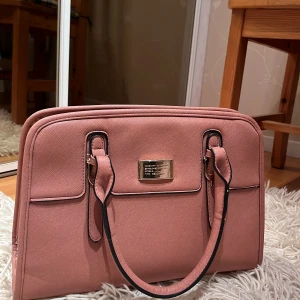 Rosa handväska från Secosana - Snygg rosa handväska från Secosana med stilren rektangulär form och guldfärgad metallplatta framtill. Väskan har två handtag med svarta detaljer, dragkedja upptill och extra fack både på utsidan och insidan. Perfekt storlek för laptop och vardagsprylar.