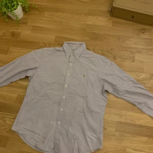 Ljusgrå/Lila skjorta från Ralph Lauren - Snygg ljusgrå skjorta från Ralph Lauren med klassisk krage och broderad logga på bröstet. Skjortan har knappar hela vägen och långa ärmar med manschett. Materialet är mjuk bomull som känns skönt mot huden. Perfekt för dig som gillar stilrena och tidlösa plagg.