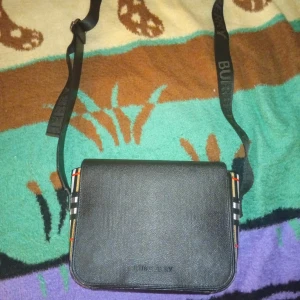 Svart axelväska från Burberry i skinn - Snygg svart axelväska från Burberry i skinn med klassiskt rutigt mönster på sidorna och justerbar axelrem med logga. Stilren design med lock och präglad Burberry-text framtill. Perfekt för dig som vill ha en ikonisk och trendig väska.