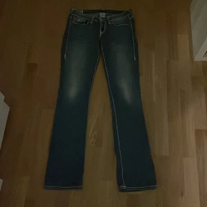 True Religion blå bootcut jeans stl 28 - Säljer ett par blå jeans från True Religion med tydliga vita sömmar och ikoniska bakfickor. Modellen har bootcut-ben och klassisk femficksdesign. Jeansen är tillverkade i USA och har en snygg tvättad look med ljusare partier. Så sjukt snyggt både till vardagen och fester, perfekt längd för mig som är 170cm! ❤️inga defekter. Har haft på mig kanske 3 gånger.