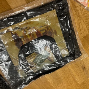 Svart shoppingväska Denim Tears x The Met - Svart shoppingväska i syntetmaterial med tryck av blommor och texten 'DENIM TEARS' samt motiv inspirerat av konst. Väskan har rektangulär form och breda handtag, perfekt för att bära med sig allt man behöver. Snygg collab mellan Denim Tears och The Met.