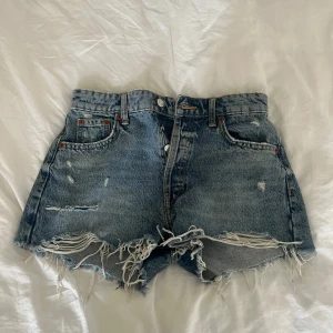 Blå högmidjade jeansshorts från Zara - Säljer ett par blå jeansshorts från Zara i storlek 34. De är högmidjade med klassisk femficksdesign, slitna detaljer och fransiga kanter för en cool vibe. Materialet är jeans med knappstängning.