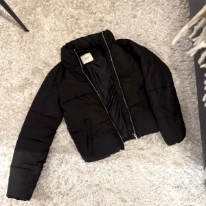 Svart pufferjacka  - Säljer en svart pufferjacka med hög krage och dragkedja framtill. Jackan är vadderad och har en clean, stilren look som passar perfekt till kalla dagar. 