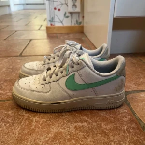 Nike Air Force 1 vit med mintgrön swoosh - Säljer ett par klassiska Nike Air Force 1 i vitt skinn med mintgrön swoosh. Skorna har perforerad tå, snörning och platt sula. Perfekta för dig som gillar en clean och sportig look med en färgklick.
