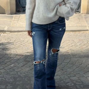 Blå bootcut jeans med slitningar - Snygga blå jeans med bootcut passform och tydliga slitningar på knäna och låren. Helt oanvända med prislappen kvar. Jeansen har vita kontrastsömmar och klassisk femficksdesign. Perfekta för en avslappnad och trendig look.