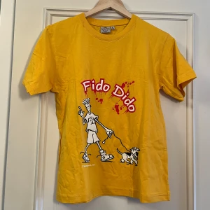 Gul Fido Dido t-shirt i bomull - Gul t-shirt från Fido Dido med coolt tryck framtill av Fido Dido och hund, samt stor röd F och logga på ryggen. Klassisk passform och rund hals. Perfekt för dig som gillar retro vibes och streetstyle.