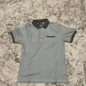Gråmönstrad piké från Salian - Snygg pikétröja från Salian i storlek S med grått och vitt geometriskt mönster. Krage och ärmslut i mörkgrönt, knappslå framtill och liten dekorativ ficka med metallmärke. Perfekt för dig som gillar stilrena detaljer och unik design.