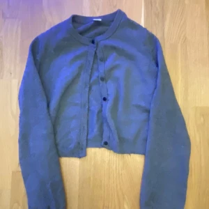 Grå croppad kofta från Lindex - Grå croppad kofta från Lindex med rund hals och svarta knappar framtill. Koftan är stickad och har långa ärmar.