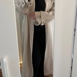 Beige lång kappa med bälte - Snygg beige kappa i lång modell med breda slag, knytskärp i midjan och stora fickor. Ärmarna har dekorativa spännen med knappar. Perfekt för dig som vill ha en stilren och trendig look under höst och vinter. Lång ända ner till ankeln.