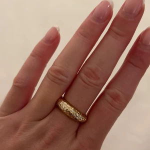 Bred guldmönstrad ring från Safira - Säljer en bred ring i guldton med snyggt ingraverat mönster som ger en cool och unik look. Ringen har en rundad form och glänsande yta som verkligen sticker ut. Perfekt för dig som vill ha ett statement-smycke till din outfit.