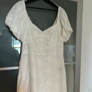 Vit broderad klänning från Nelly - Superfin vit kortklänning från Nelly med broderade blommor, puffärmar och v-ringning. Klänningen har knappar framtill, volangkant nertill och är tillverkad i ett lätt bomullstyg. Perfekt för sommarens alla tillfällen! Helt oanvänd