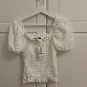 Vit blus med puffärmar från Gina Tricot  - Supersöt vit blus från Gina Tricot med smockad kropp, puffiga korta ärmar och volangkant nertill. Blusen har broderade detaljer och en fyrkantig urringning som ger en romantisk vibe. Perfekt för dig som gillar en feminin och trendig stil.