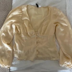 Gul blus med knappar - Snygg gul blus i glansigt satinliknande material med v-ringning och puffiga långärmar. Blusen har knappar framtill och en figurnära passform som ger en romantisk vibe. Perfekt att styla med jeans eller kjol.