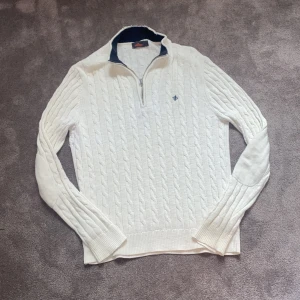 Vit kabelstickad half zip-tröja från Morris - Snygg vit stickad tröja från Morris med kabelstickat mönster och half zip-dragkedja. Tröjan har lång ärm, ribbade muddar och en broderad logga på bröstet. Perfekt för lager-på-lager och chillare dagar. Hör av dig vid funderingar så svarar jag direkt! Nypris uppåt 2000 kr