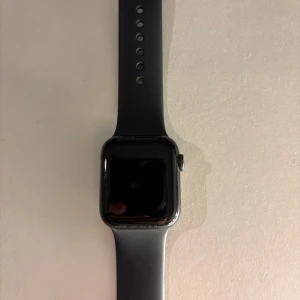 Apple Watch SE svart 40mm  - Apple Watch SE 40mm i svart med stilren, rundad fyrkantig urtavla och svart silikonarmband. Klockan har digital krona på sidan och kommer med original laddkabel. Perfekt för dig som gillar smarta funktioner och snygg design.