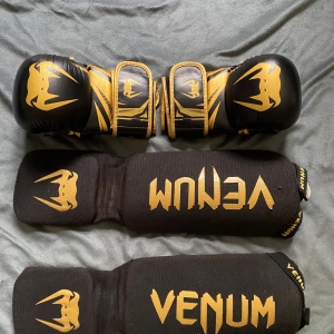 Svarta och guld MMA-handskar Venum - Säljer ett par svarta och guldiga MMA-handskar från Venum med matchande benskydd. Handskarna har öppna fingrar, vaddering och justerbara kardborreband. Benskydden är svarta med guldigt Venum-tryck och elastiska band för bra passform. Perfekt för kampsportsträning.