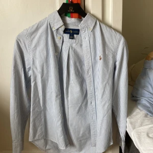 Randig blåvit skjorta från Ralph Lauren - Snygg blåvit randig skjorta från Ralph Lauren med klassisk button-down krage och broderad logga på bröstet. Skjortan har långa ärmar, knappar framtill och är tillverkad i mjuk bomull. Perfekt för en clean och stilren look. Skick 9.5/10, använd kanske 1 gång. Strlk 14