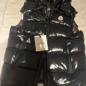 Svart dunväst från Moncler - Snygg svart dunväst från Moncler med glansig finish och klassisk logga på bröstet. Västen har två dragkedjeförsedda fickor framtill och quiltad design. Insidan har ett coolt serietryck och Moncler-märke. Perfekt för lager-på-lager under höst och vinter. Västen är storlek 1=S
