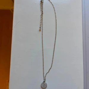 Silverfärgat halsband med glittrig rund berlock - Snyggt halsband i silverfärg med en rund berlock som är täckt av små glittrande stenar. Kedjan är tunn och har justerbar längd med förlängningskedja. 