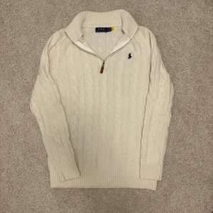 Vit kabelstickad tröja Polo Ralph Lauren - Kabelstickad vit tröja från Polo Ralph Lauren med halv dragkedja och hög krage. Klassisk design med broderad logga på bröstet och ribbade muddar. Perfekt för lager-på-lager och snygg till jeans eller chinos.Passar L/Xl