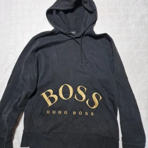 Svart hoodie från Hugo Boss med guldtryck - Svart hoodie från Hugo Boss med stort guldigt BOSS-tryck framtill. Klassisk huva med snörning och långa ärmar. Perfekt för dig som vill ha en stilren och lyxig streetwear-look. Sälj ej längre. 