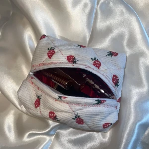 Handgjord sminkväska med jordgubbar - Supergullig liten handgjord sminkväska i vit manchester med quiltad struktur och sött jordgubbsmönster. Väskan har dragkedja upptill och är perfekt för att bära småsaker. Perfekt accessoar för dig som gillar unika detaljer och lekfull stil.