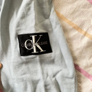 Ljusgrå hoodie från Calvin Klein Jeans - Snygg ljusgrå hoodie från Calvin Klein Jeans med stor CK-logga på ärmen. Tröjan har huva med snörning och en klassisk känguruficka framtill. Perfekt för en chill och stilren look. Den är nytvättad och behöver strykas 