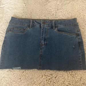 Blå jeanskjol från Vero Moda - Snygg blå jeanskjol från Vero Moda med klassisk femficksdesign, bälteshällor och rå kant nertill. Kjolen har en rak passform och är tillverkad i slitstarkt denimtyg. Perfekt för dig som gillar en enkel och tidlös look.