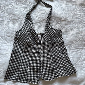 Rutig svartvit halterneck topp - Supersöt svartvit rutig korsettopp från H&M med halterneck och knyt i nacken. Toppen har hakknäppning framtill och smock baktill för extra komfort. Storlek 38 men passar mer som S
