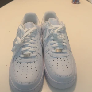 Nike Air Force 1 helvita sneakers - Klassiska Nike Air Force 1 sneakers i helvitt skinn med perforerad tå, snörning och ikonisk swoosh på sidan. Platt sula och låg modell som passar perfekt till streetwear. Metallbricka med 'AF1' på snörningen ger extra detalj. Storlek 44.5 box ingår