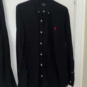 Svart skjorta från Polo Ralph Lauren - Svart långärmad skjorta från Polo Ralph Lauren i slim fit-modell. Skjortan har klassisk krage, vita knappar framtill och röd broderad logga på bröstet. Tillverkad i mjuk bomull som känns skön mot huden. Perfekt för dig som gillar stilrena och tidlösa plagg.