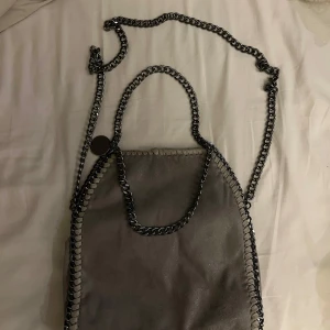 Grå axelväska med kedjedetaljer - Snygg grå axelväska från Stella McCartney med grova silverfärgade kedjor som handtag och axelrem. Väskan är i mjuk mocka och har ett coolt leopardmönstrat innerfoder. Perfekt för dig som gillar statement-accessoarer.