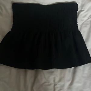 Svart bandeau tubtopp - Svart bandeau tubtopp med smockad överdel och utsvängd nederdel. Toppen är axelbandslös och har en enkel, stilren look som passar perfekt till sommarens outfits.