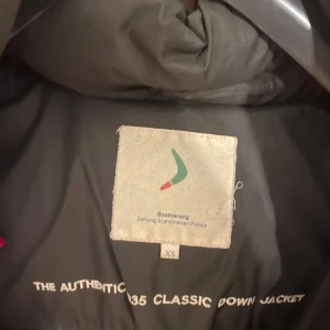 Brun dunjacka från Boomerang XS - Säljer en brun dunjacka från Boomerang i klassisk modell. Jackan har hög krage, dragkedja och knappar framtill samt broderad logga på ärmen. Fodrad och riktigt varm, perfekt för kalla dagar. Snygg och tidlös design som funkar till det mesta.