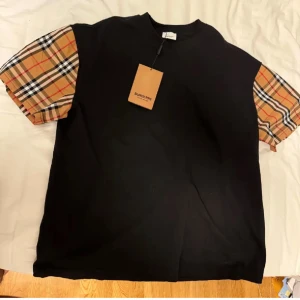 Burberry t-shirt med rutiga ärmar - Svart t-shirt från Burberry med klassiska rutiga kortärmar i beige, rött och svart. Oversized passform och rund halsringning. Perfekt statement-plagg för dig som gillar lyxiga streetwear-vibbar.