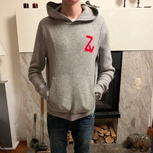 Grå hoodie med röd döskalle från Zadig voltair  - Grå stickad hoodie med stor röd döskalle på ryggen och röd Z-logga på bröstet. Tröjan har huva, magficka och ribbade muddar. Perfekt för dig som gillar edgy och unik stil.