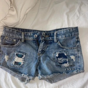 Lågmidjade blå jeansshorts med slitningar och nitar - Jättefina och unika jeans med slitningar och nitar, aldrig använda men har ingen prislapp. Köpta i Spanien ❤️. Säljs pga att de är lite för små för mig som brukar ha 36, dessa är mer åt S hållet. 