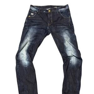 G-Star Raw mörkblå jeans med slitningar - Snygga mörkblå jeans från G-Star Raw med coola slitningar och kontrastsömmar. Klassisk femficksmodell med logga på bakfickan och metallknappar. Jeansen har en slim/straight passform och är tillverkade i robust denim.                                 W32 L34 Pris kan diskuteras 