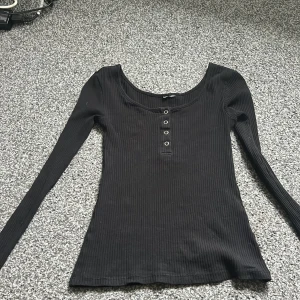 Svart ribbad långärmad topp med knappar - Svart ribbad långärmad topp med rundad halsringning och fyra dekorativa metallknappar framtill. Figurnära passform och stretchigt material som känns skönt mot kroppen. Perfekt att styla med jeans eller kjol för en clean look.