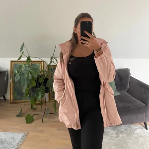 Ljusrosa parkajacka med huva - Säljer en ljusrosa parkajacka med huva och dragsko i midjan. Jackan har både dragkedja och knappar framtill, samt fickor med lock. Perfekt för dig som vill ha en snygg och bekväm jacka till vår och höst.