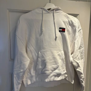 Vit hoodie från Tommy Jeans - Vit hoodie från Tommy Jeans med broderad logga på bröstet. Klassisk känguruficka framtill, dragsko i huvan och ribbade muddar. Perfekt för en chill och stilren look. Materialet är mjuk bomull som känns skönt mot huden.