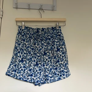 Blommiga shorts från Lindex i blått - Snygga shorts från Lindex med blått och vitt blommönster. De har elastisk midja och är tillverkade i ett mjukt, lätt tyg som passar perfekt för varma dagar. Shortsen har en avslappnad passform och är superbekväma att bära.